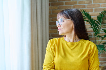 Middle-aged woman looking out the window, calm thoughtful female in yellowの写真素材
