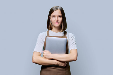 Portrait of young woman in apron holding laptop on gray background