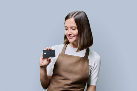 Young happy woman service worker in apron with credit card in hand, on gray backgroundの写真素材