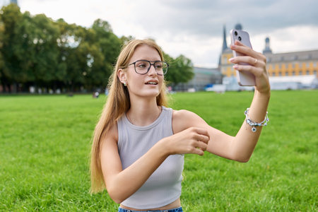 Teenage girl student having video call on smartphone, sitting on grass near universityの写真素材