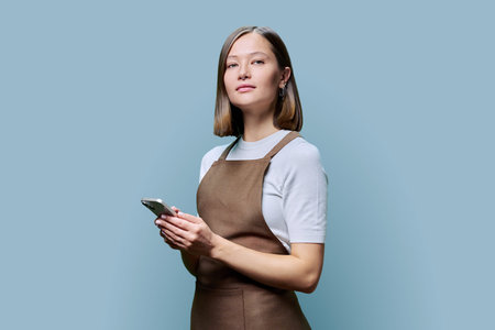 Portrait of young woman in apron holding smartphone in hands on blue background. Serious female...