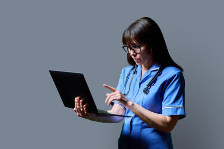 Female nurse using laptop, profile view on gray studio backgroundの写真素材