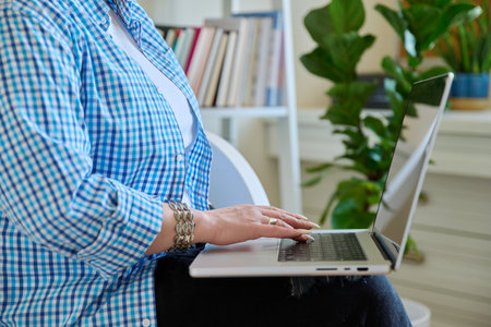 Close-up of mature womans hands typing on laptop, home interior backgroundの写真素材