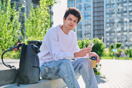 Relaxed young male with smartphone, glass of coffee looking at camera, outdoorの写真素材