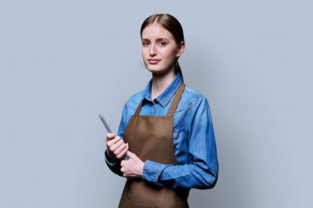 Young female worker in apron holding digital tablet on gray studio backgroundの写真素材