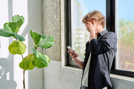 Upset sad teenager guy looking at smartphone screen, at home near windowの写真素材