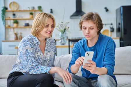 Middle aged mother and teenage son sitting together at home looking at smartphoneの写真素材