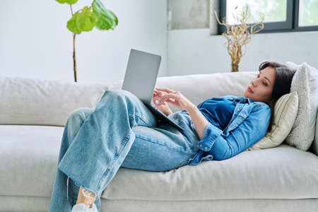 Young serious woman typing on laptop lying on couch at homeの写真素材