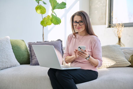 Middle aged woman sitting on sofa with laptop smartphone, in living room interior. Mature female usingの写真素材