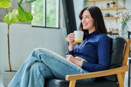 Portrait of beautiful 30s smiling woman with cup sitting in armchair at home in living roomの写真素材