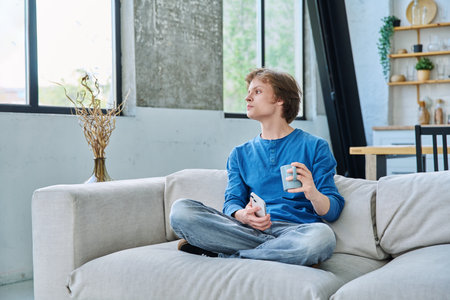 Relaxed teenager guy sitting at home on the couch with smartphone and cup of teaの写真素材