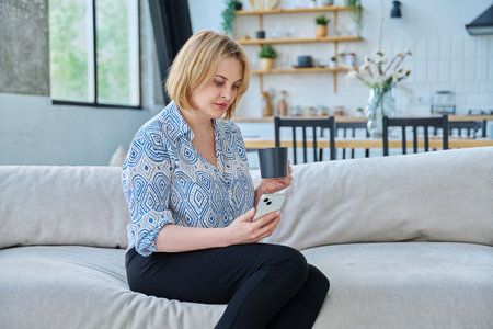 Portrait of middle aged blonde woman with smartphone cup sitting on sofa in living roomの写真素材