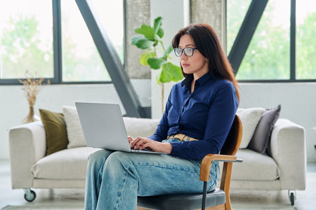 Portrait of serious business woman typing on laptop in office with couchの写真素材