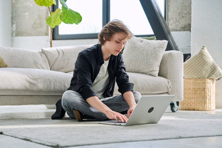 Handsome serious teenage boy sitting at home on the floor typing on laptopの写真素材