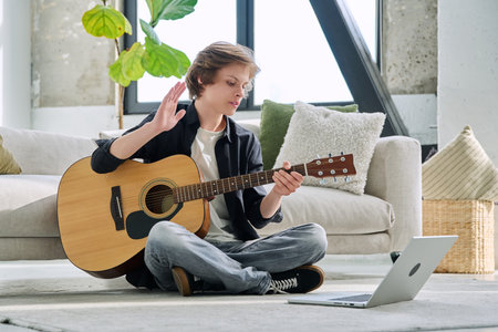 Teenage boy with acoustic guitar and laptop, having online music lessonの写真素材