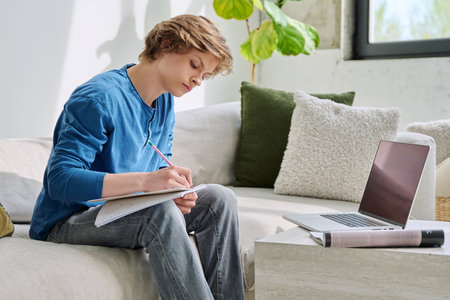 Teenager guy student studying sitting at home on sofa using laptop textbook notebookの写真素材