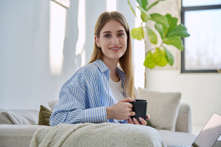 Portrait of young beautiful smiling blonde woman with cup of drink sitting on sofa at homeの写真素材