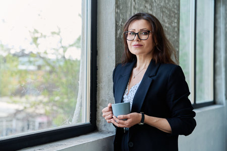 Portrait of middle aged beautiful businesswoman looking at camera, standing near windowの写真素材