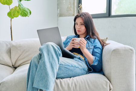 Young relaxed woman sitting on sofa at home with cup, looking at laptopの写真素材