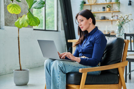 Young serious woman typing on laptop sitting on chair at homeの写真素材