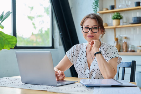 Middle aged smiling woman working with laptop, looking at camera, sitting at homeの写真素材
