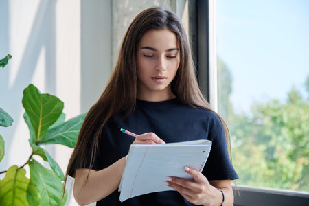 Portrait of teenage college student girl with notebook pencil at home near window