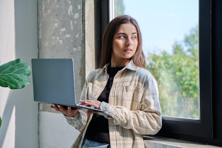 Portrait of young teenage girl using laptop standing at home, looking at windowの写真素材