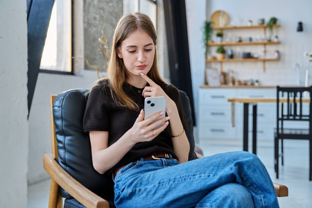 Young woman sitting at home on chair using smartphoneの写真素材