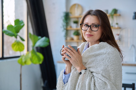 Relaxed middle aged woman with mug under blanket in home interiorの写真素材