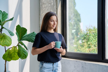 Portrait of beautiful calm relaxed teenage girl with cup in hands at home looking out windowの写真素材