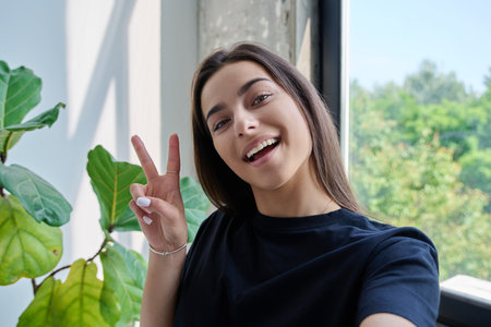 Close-up selfie portrait of teenage female looking at web camera showing victory gestureの写真素材
