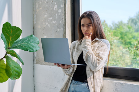 Portrait of serious teenage girl standing with laptop in home interior near windowの写真素材