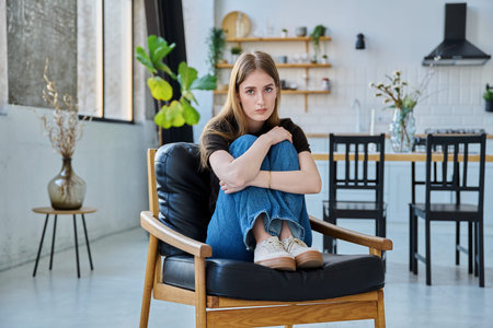 Portrait of young beautiful serious woman looking at camera sitting at home in chairの写真素材
