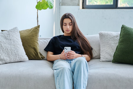 Young serious woman sitting at home on couch using smartphoneの写真素材