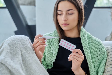 Sad upset teenage girl looking at thermometer holding blister pack of pills sitting at homeの写真素材
