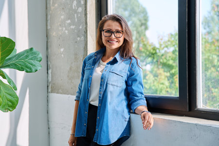 Middle aged beautiful smiling woman looking at camera standing near windowの写真素材