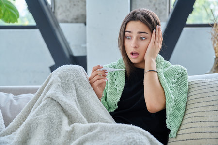 Sad surprised teenage girl looking at thermometer sitting at home under blanketの写真素材