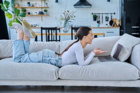 Young teenage girl using laptop for video chat lying on sofa at homeの写真素材