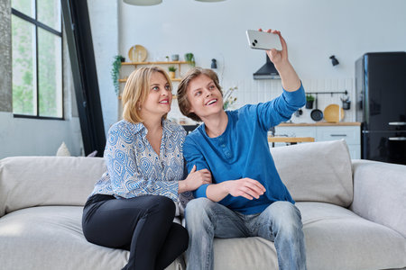 Middle aged mother and teenage son sitting together at home taking selfie photo on smartphoneの写真素材