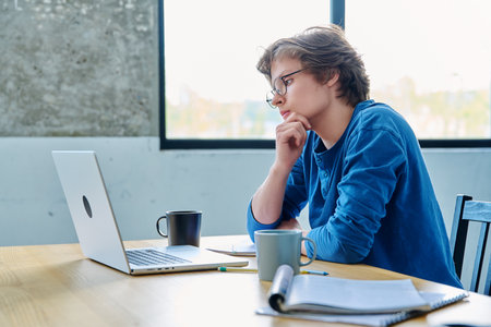 Teenage guy student sitting at the table with laptop, textbooks in home interiorの写真素材