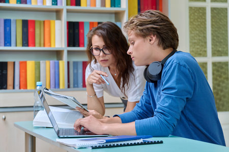 Teenage student guy studying with female teacher inside classroom libraryの写真素材