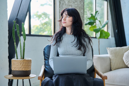Portrait of young woman sitting in chair typing on laptop, in home interior