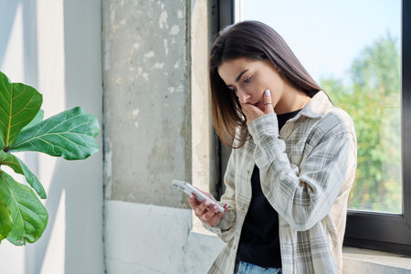 Serious teenage female standing at home reading texting on smartphoneの写真素材