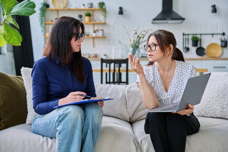 Female social worker interviewing middle aged woman, sitting together in living roomの写真素材