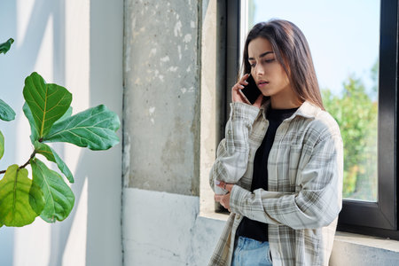 Serious teenage female standing at home talking on smartphoneの写真素材