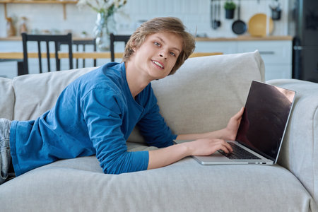 Handsome teenage boy lying at home on couch with laptop, looking at cameraの写真素材
