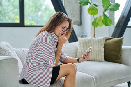 Sad serious middle aged woman with smartphone sitting on sofaの写真素材
