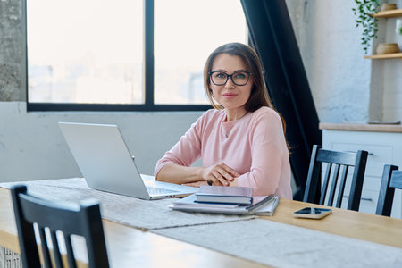 Middle aged smiling woman working with laptop, looking at camera, sitting at homeの写真素材