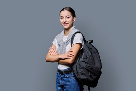 Portrait of confident female teenage student with backpack on gray background