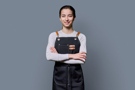 Portrait of young smiling female student in apron on gray background